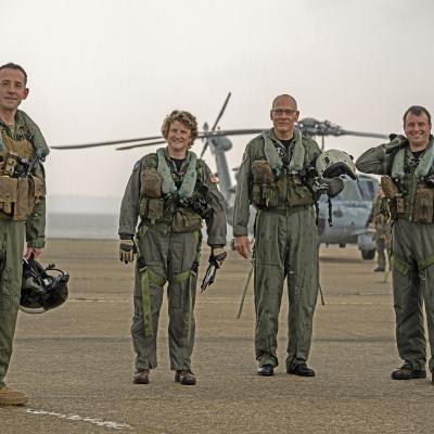 Aviators in flight suits stand in front of a helicopter on a flight line against a smoky horizon on the Patuxent River.