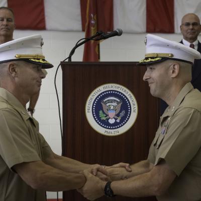 Two marines shake hands after a change of command ceremony in front of a podium with the PMA-274 seal