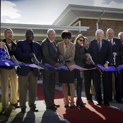 Professionals stand shoulder-to-shoulder and cut a blue ribbon founding a new test facility under sunny skies.