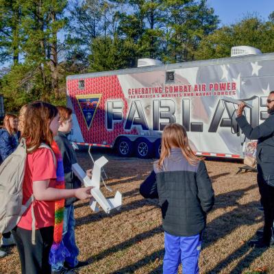Man on the left stands in front of the FABLAB trailer holding a foam glider airplane and children stand on the left watching him