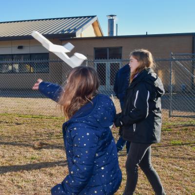 Two female students stand outside and one is throwing a foam airplane into the air