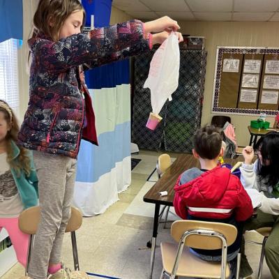 Student stands on top of chair holding a project made with a plastic bag 