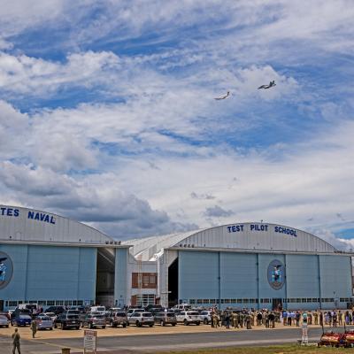 A C-12 Huron fixed-wing aircraft flies ahead of an F/A-18 Super Hornet over school hangars above a crowd of event attendees on the flight line.
