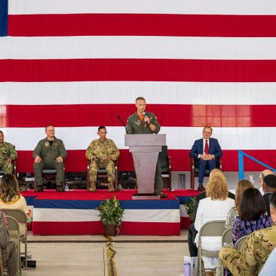 A military aviator stands behind a podium delivering remarks in on a stage in hangar spaces.