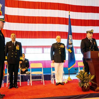 Military members stand on a stage with a large American flag as backdrop during a change of command ceremony