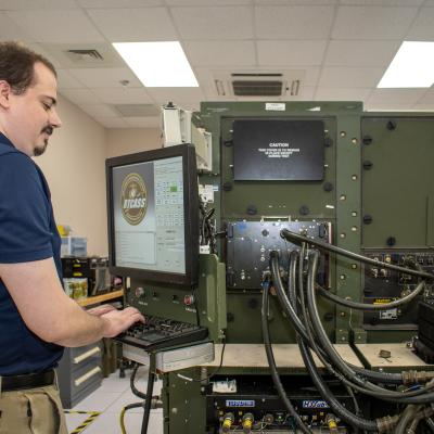 Man in blue shirt works at computer in front of large, green machine 