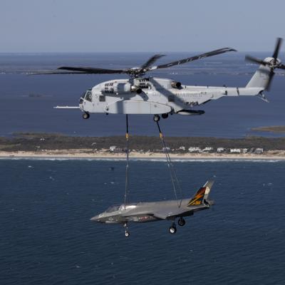 A helicopter carries a F-35 airplane by straps across open water