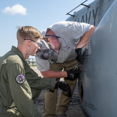 Marine and man in grey shirt work on exterior of military aircraft 