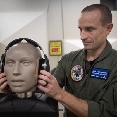 An aeromedical officer fits a dummy with headphones for audio testing in a lab.