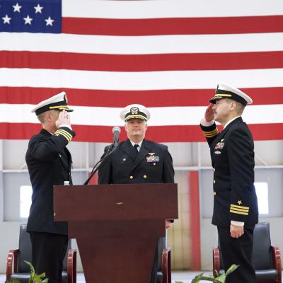 Naval officers salute each other in front of a podium during a ceremony inside a hangar.