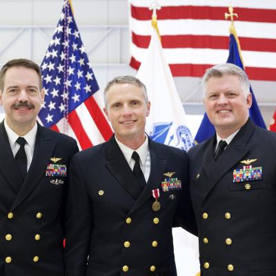 Three naval officers gather for a photo in front of flags inside a hangar space.