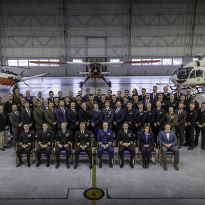 Four rows of people pose for a picture in front of five aircraft. The first row is seated, the back three rows are standing. Most are wearing their military dress uniform. There are a few civilians in business attire.