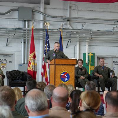 A military officer speaks to an audience from a podium in an aircraft hangar.