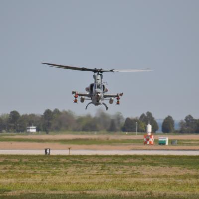 Helicopter hovering over a green lawn and part of a runway with trees in the background