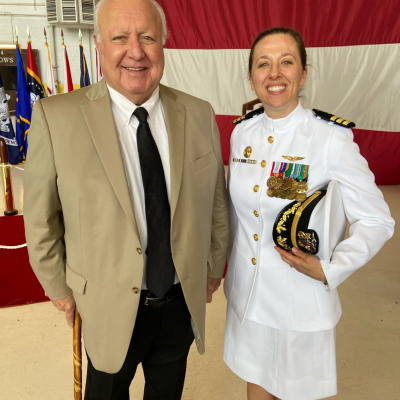 A man in a khaki blazer standing with a woman naval officer in Navy dress whites
