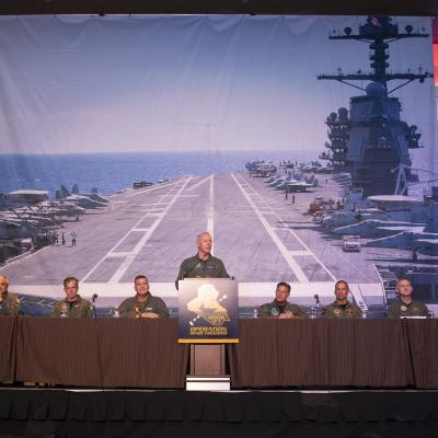 Man in flight suit stands at podium, men in flight suits sit at long table, photo of aircraft carrier deck behind them.