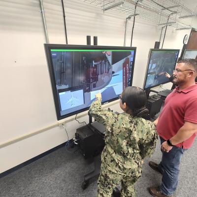 Sailor in uniform conducts training on a virtual touch-screen while being supervised by an instructor in a red shirt