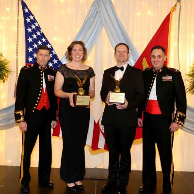 Four people standing on stage with awards and the USA and USMC flags in the background