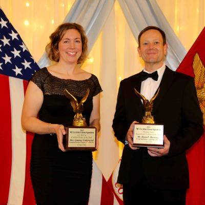 Man and woman standing on stage with awards and the USA and USMC flags in background