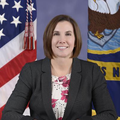 Official photo of a woman in a blazer standing in front of the American and Navy flags