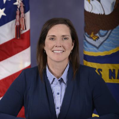 Woman with brown hair wearing a Navy blazer and standing in front of the American and Navy flags