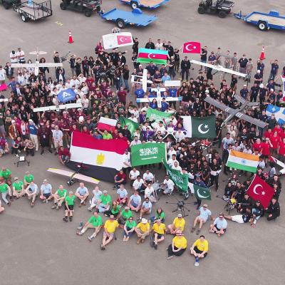 Dozens of students gather for a photo on a flight line posing with small unmanned systems and their home country flags.