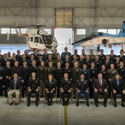 Multiple developmental test professionals pose for an official class portrait in a hangar with aircraft stationed in the background.