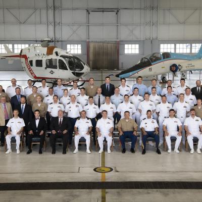 Military and civilian test pilots in uniform and formal attire pose for a class photo inside a hangar with a helicopter and jet. 