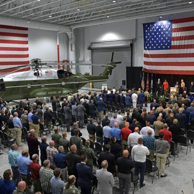 People seated in rows with a stage in front and a helicopter and two American flags 