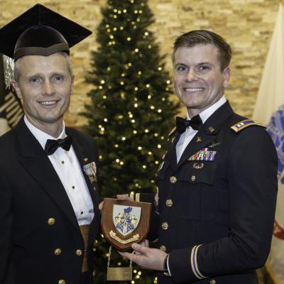 A Navy officer and an Army Officer pose in front of flags and a Christmas Tree. the Army officer is holding an award plaque. 