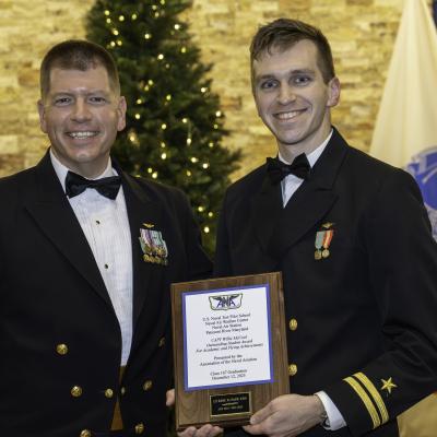 Two Naval Officers pose in front of flags. The one on the left is holding a plaque.
