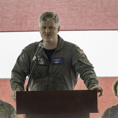 The Naval Test Wing Atlantic Commodore addresses ceremony attendees in a hangar at a podium.