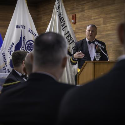 With the backs of attendee's heads in the foreground, a Navy Rear Admiral Speaks at a wooden podium in front of three flags. 