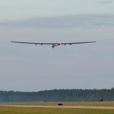 A large unmanned aerial vehicle is flying low to the ground above a runway. There are trees in the background.