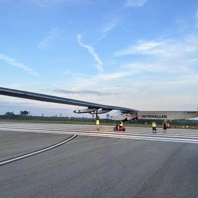A large unmanned aerial system is pictured on a runway prior to takeoff. There are four people in the picture who are preparing the UAS for takeoff.