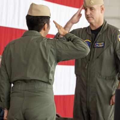 Two naval officers exchange salutes in front of a large American flag.