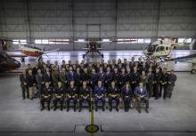 Four rows of people pose for a picture in front of five aircraft. The first row is seated, the back three rows are standing. Most are wearing their military dress uniform. There are a few civilians in business attire.