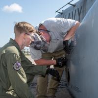 Marine and man in grey shirt work on exterior of military aircraft 