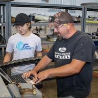 Male aircraft work leader demonstrates use of computer-based maintenance manual for high-school aged, female summer intern. 