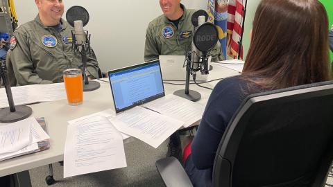Two pilots in flight suits sitting at a table with microphones recording a podcast.