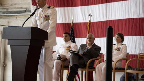 An officer in a white Navy dress uniform speaks behind a podium against an American flag backdrop.