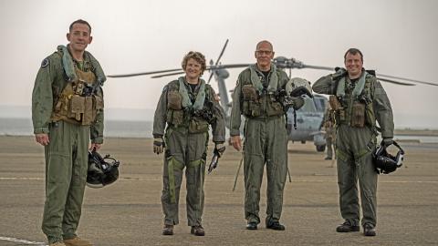 Aviators in flight suits stand in front of a helicopter on a flight line against a smoky horizon on the Patuxent River.