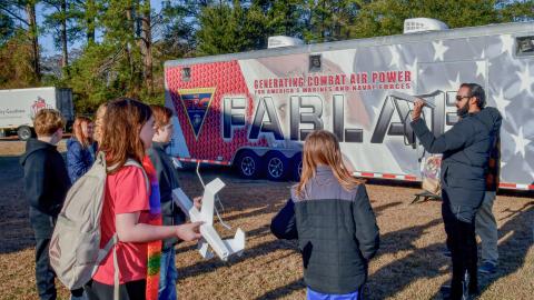 Man on the left stands in front of the FABLAB trailer holding a foam glider airplane and children stand on the left watching him