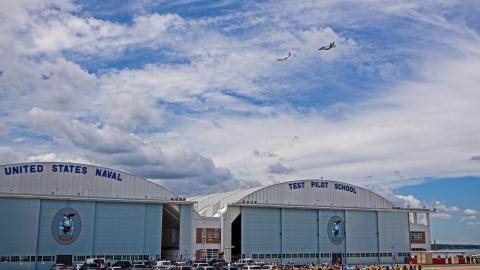 A C-12 Huron fixed-wing aircraft flies ahead of an F/A-18 Super Hornet over school hangars above a crowd of event attendees on the flight line.