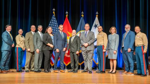 group of business and military professionals standing in a line on stage with flags behind them