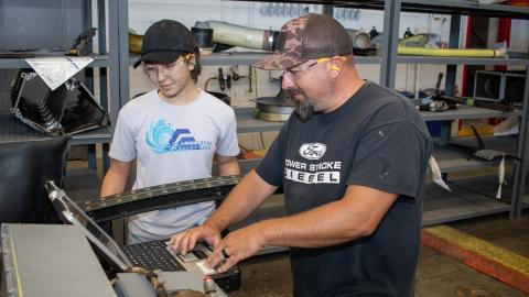 Male aircraft work leader demonstrates use of computer-based maintenance manual for high-school aged, female summer intern. 