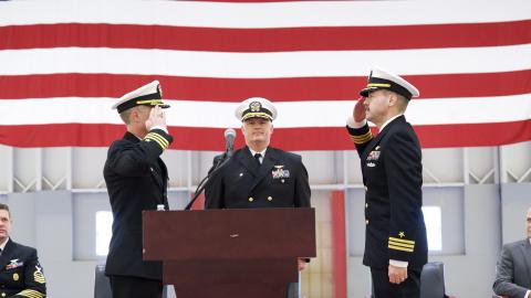 Naval officers salute each other in front of a podium during a ceremony inside a hangar.