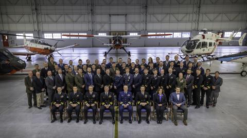 Four rows of people pose for a picture in front of five aircraft. The first row is seated, the back three rows are standing. Most are wearing their military dress uniform. There are a few civilians in business attire.