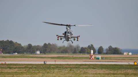 Helicopter hovering over a green lawn and part of a runway with trees in the background