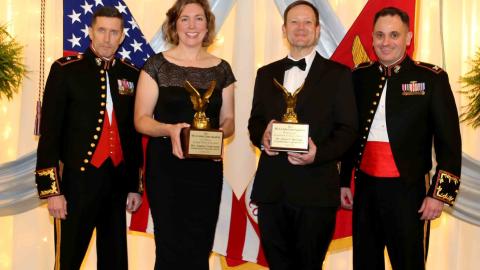 Four people standing on stage with awards and the USA and USMC flags in the background